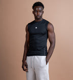 A man stands against a solid warm tan studio background wearing a black sleeveless athletic shirt and light beige sweatpants.