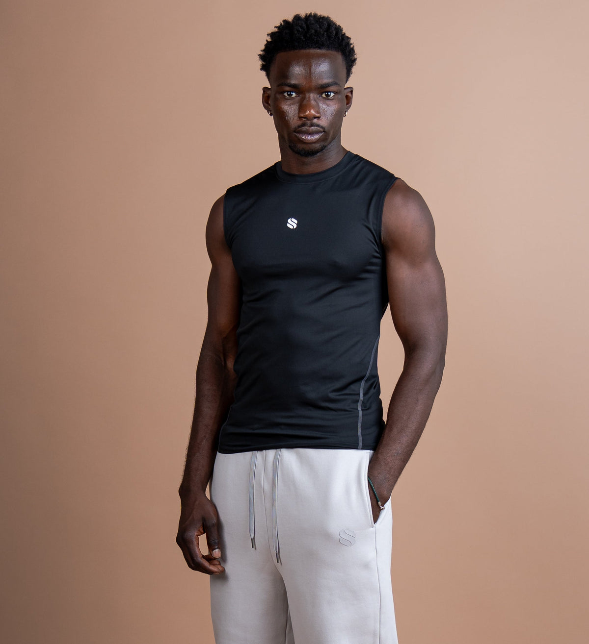 A man stands against a solid warm tan studio background wearing a black sleeveless athletic shirt and light beige sweatpants.