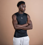 A man wearing a black sleeveless athletic tank top and white sweatpants, standing with arms crossed against a solid warm beige background.