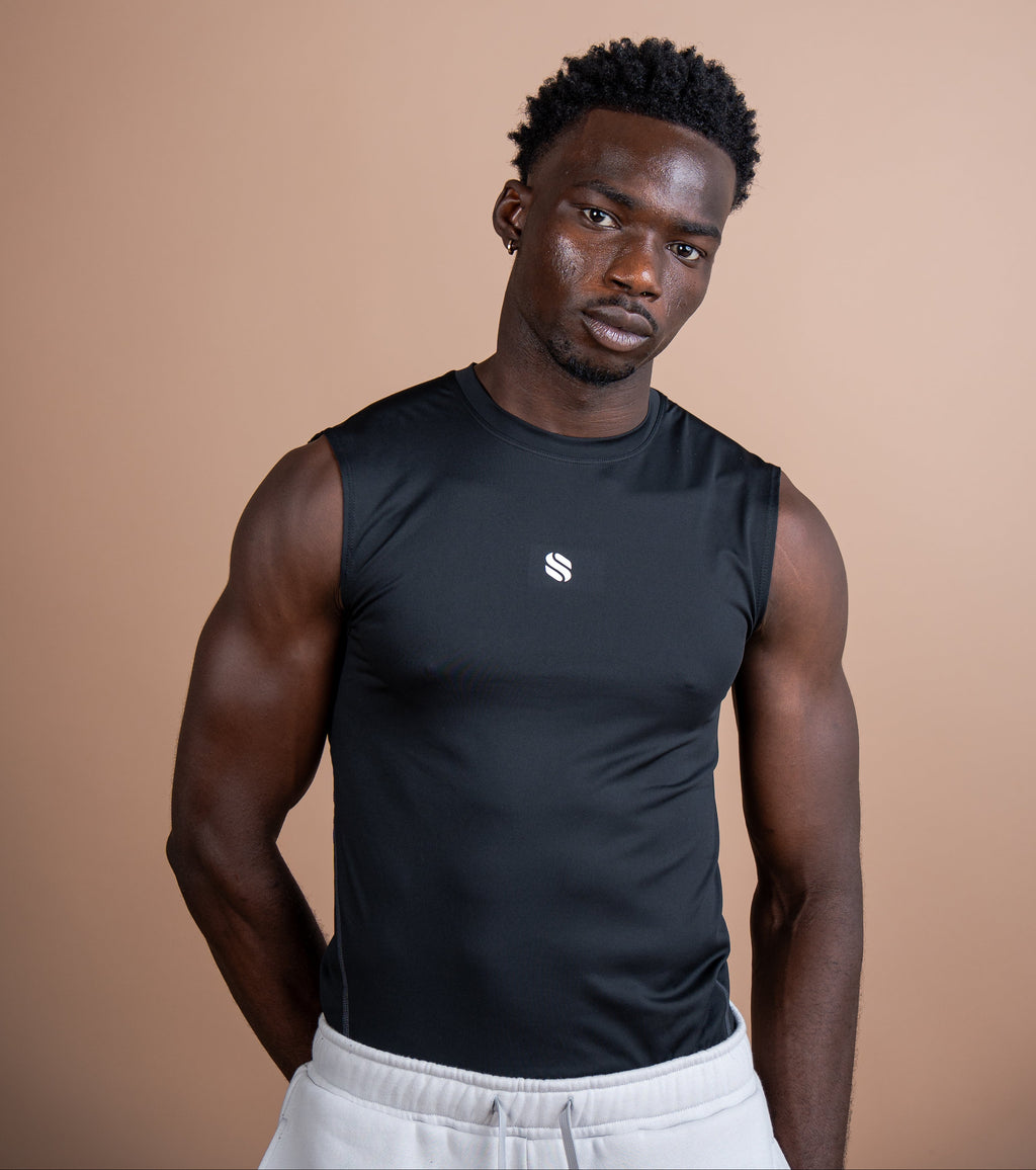 A man wearing a black sleeveless athletic tank top with a small white logo on the chest, paired with light gray sweatpants, standing against a solid warm beige background.