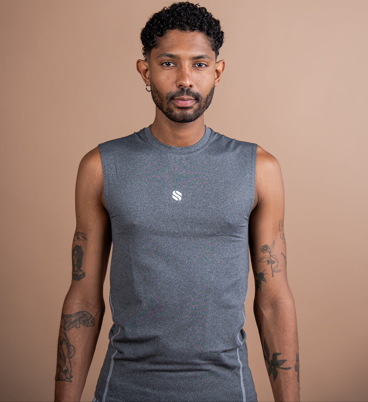 A man wearing a sleeveless heather gray athletic shirt with a small white logo on the chest, standing against a solid tan background.