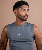 A man with short curly black hair and a beard wearing a gray sleeveless athletic shirt with a small white logo on the chest, photographed against a solid warm beige background.