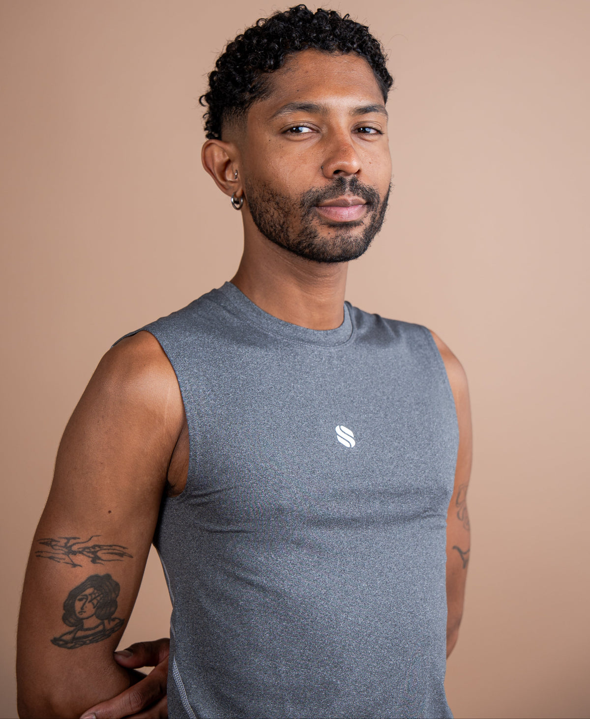 A man with short curly black hair and a beard wearing a gray sleeveless athletic tank top with a small white logo on the chest, standing against a solid warm beige background.