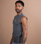A man wearing a dark gray sleeveless athletic tank top and light gray shorts, standing against a solid warm tan background.