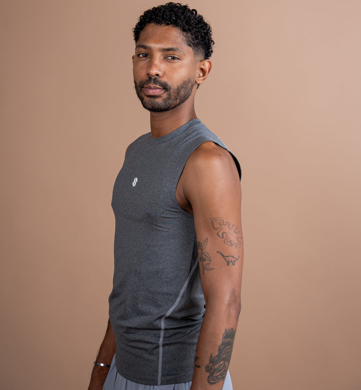 A man wearing a dark gray sleeveless athletic tank top and light gray shorts, standing against a solid warm tan background.