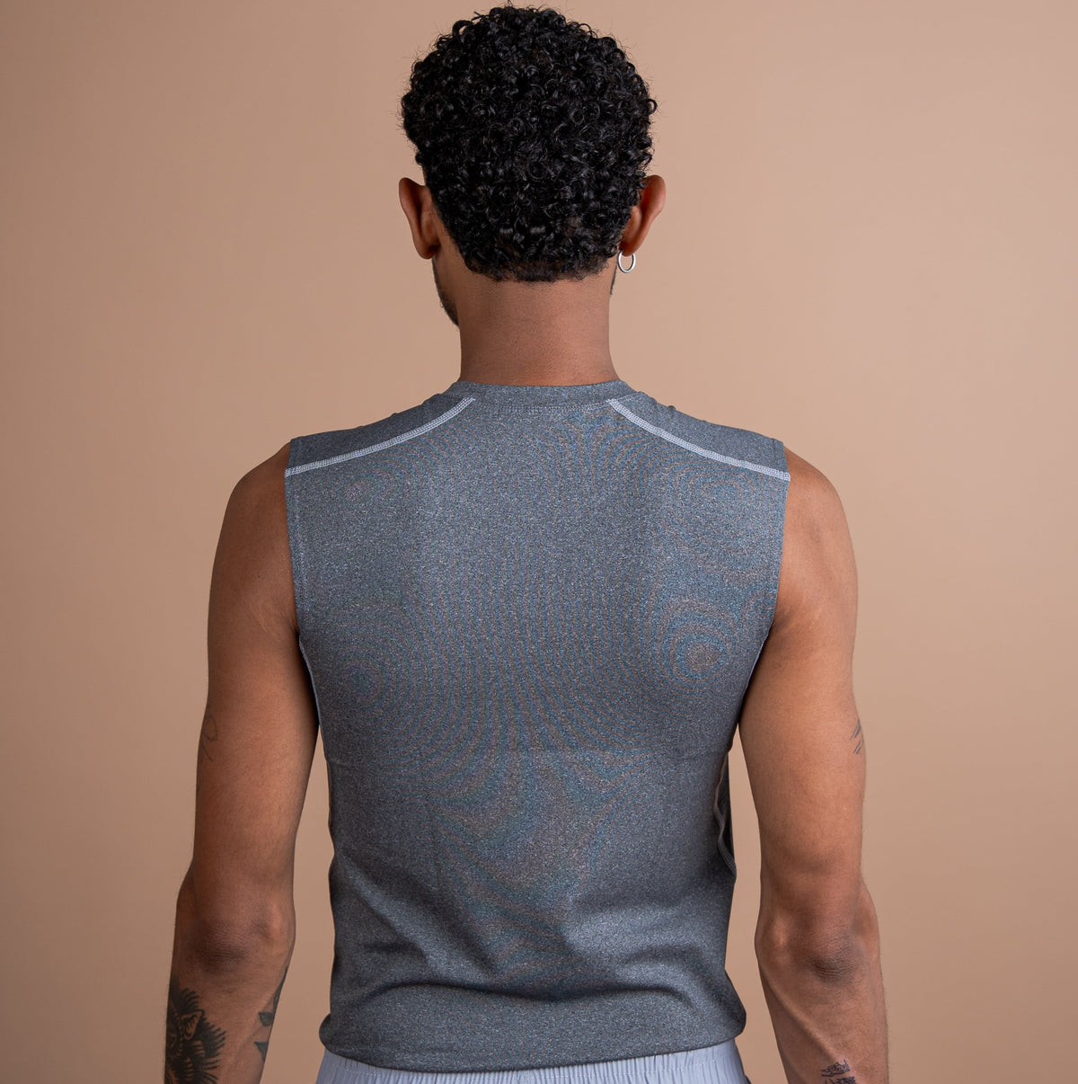 A man viewed from behind wearing a sleeveless gray athletic tank top and light gray shorts against a solid beige studio background.
