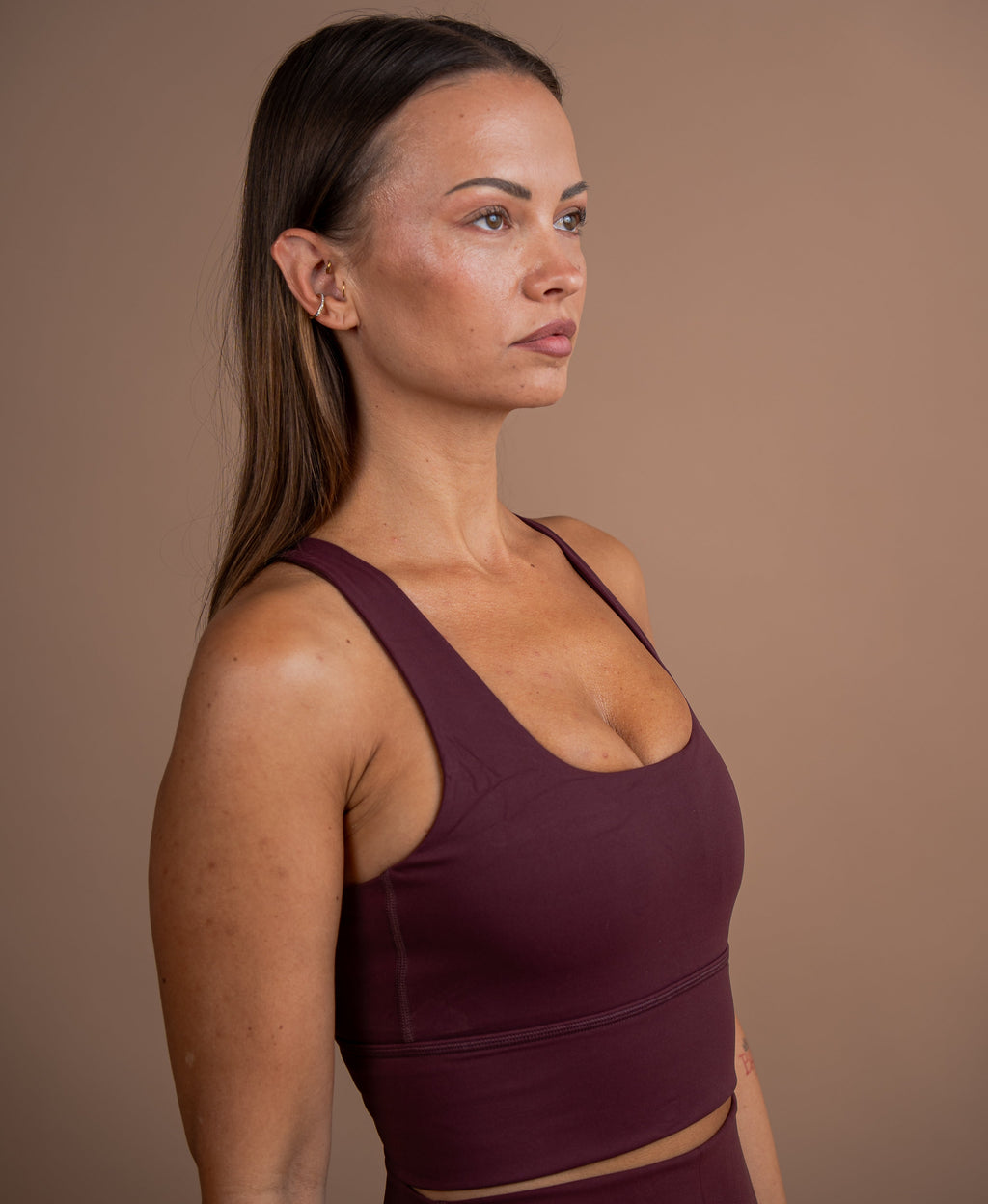 A woman in profile wearing a dark plum purple sports bra and matching leggings against a solid taupe background.