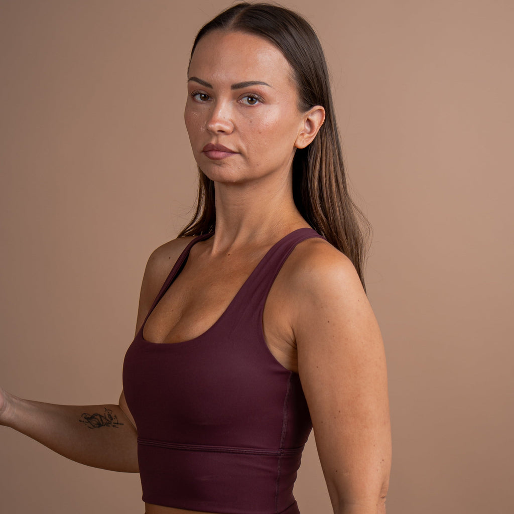 A woman modeling a matching burgundy activewear set, featuring a scoop-neck crop top and high-waisted leggings, set against a solid warm brown studio background.