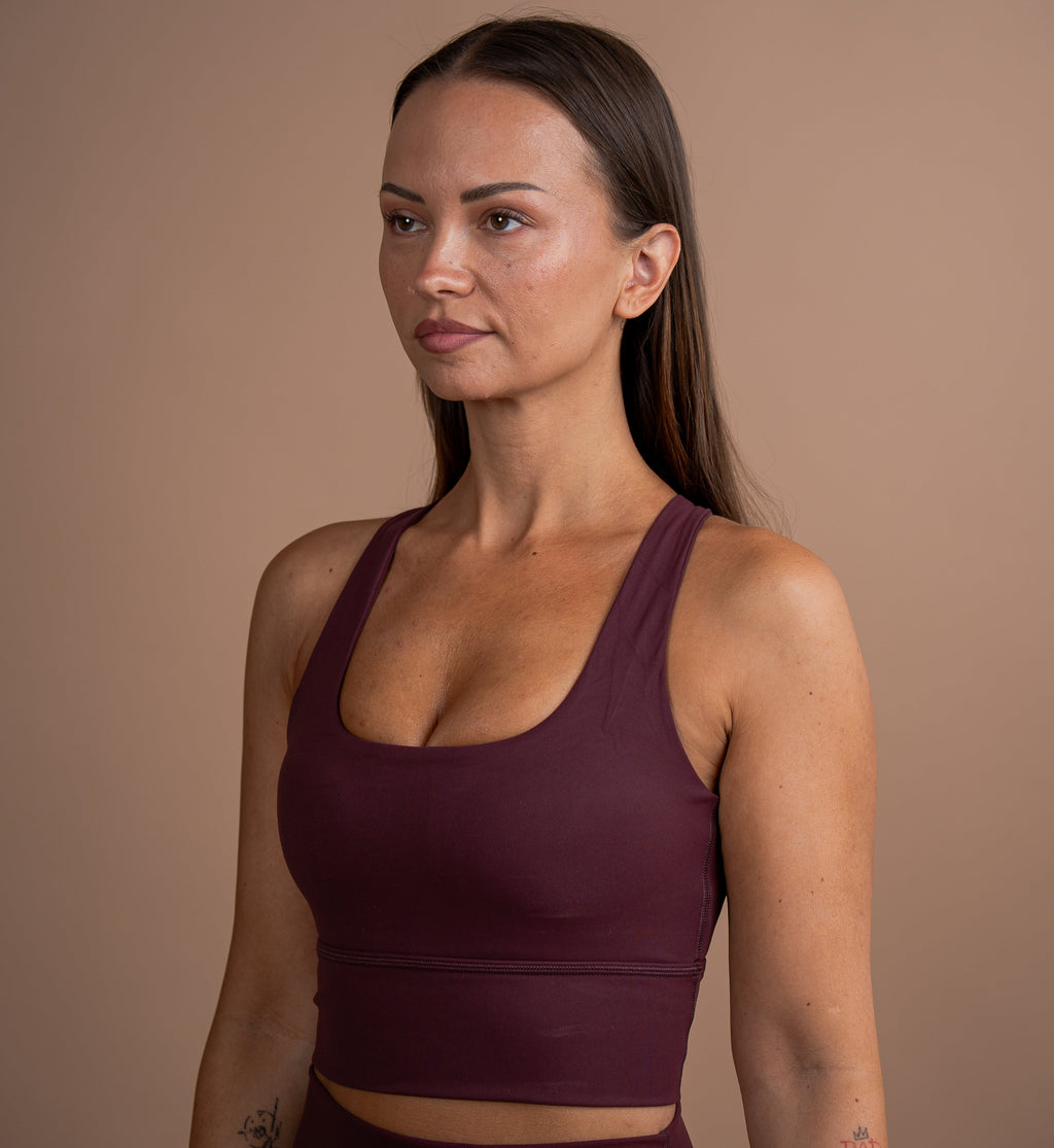 A woman with long dark hair wearing a matching dark plum purple sports bra and high-waisted leggings set, standing against a solid warm brown studio background.