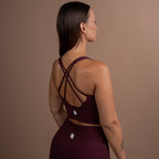 A woman shown from the back wearing a matching burgundy activewear set, featuring a sports bra with crisscross straps and high-waisted leggings, set against a solid taupe background.