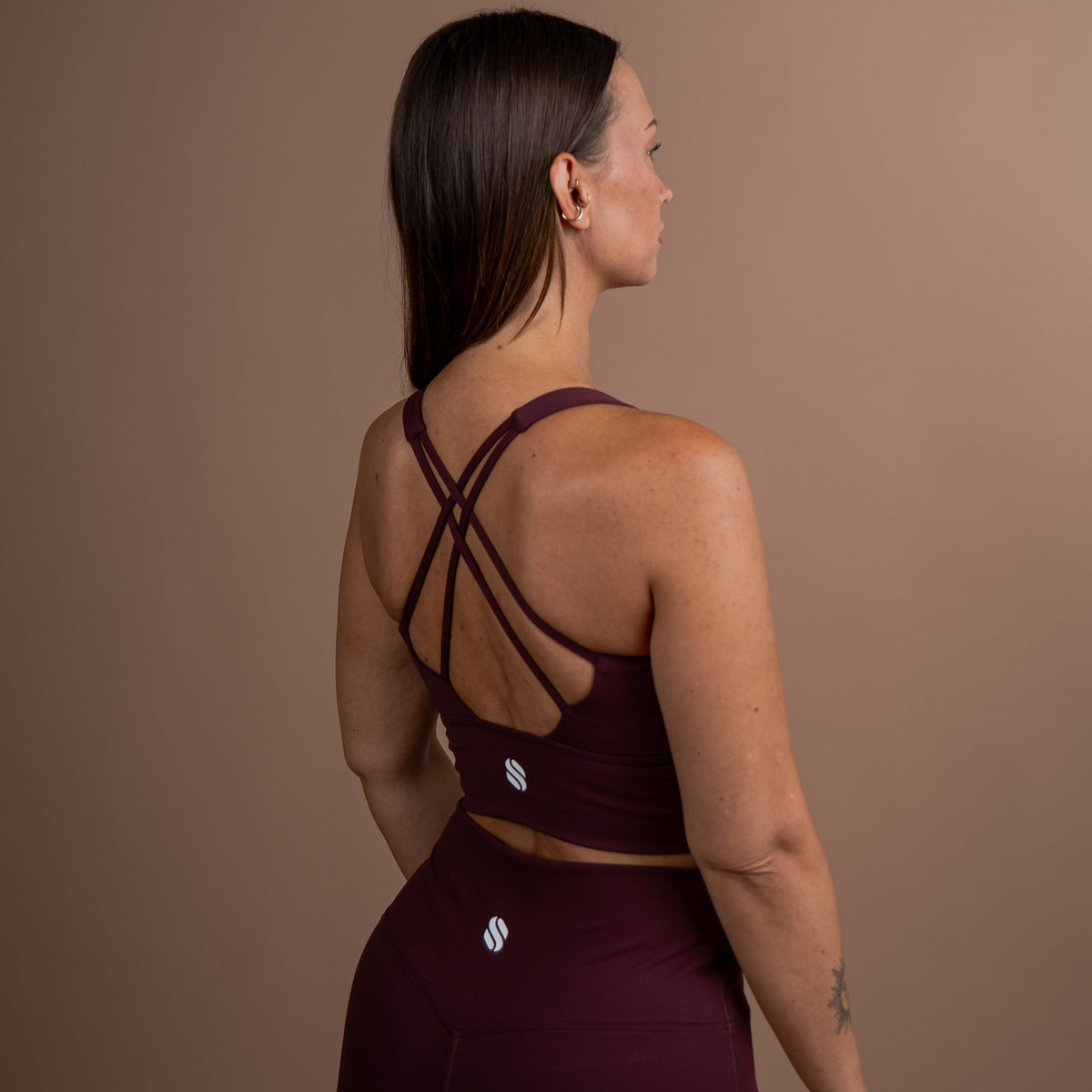 A woman shown from the back wearing a matching burgundy activewear set, featuring a sports bra with crisscross straps and high-waisted leggings, set against a solid taupe background.
