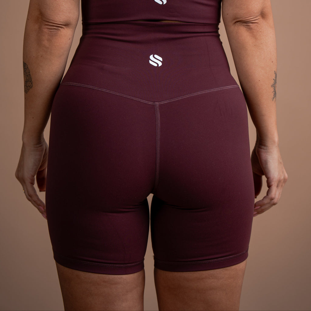 Rear view of a woman wearing a matching burgundy activewear set with high-waisted shorts and a strappy top, featuring a small white stylized 'S' logo on the back of both pieces, set against a solid warm brown background.