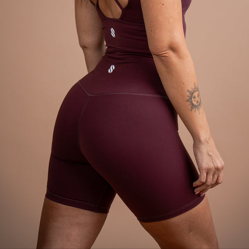 A woman wearing a matching burgundy athletic set consisting of a racerback tank top and high-waisted biker shorts, shown from a rear three-quarter angle against a solid beige studio background.