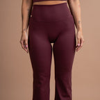 A model wearing a matching burgundy activewear set consisting of a scoop-neck crop top and high-waisted flared leggings, shown against a solid warm beige studio background.