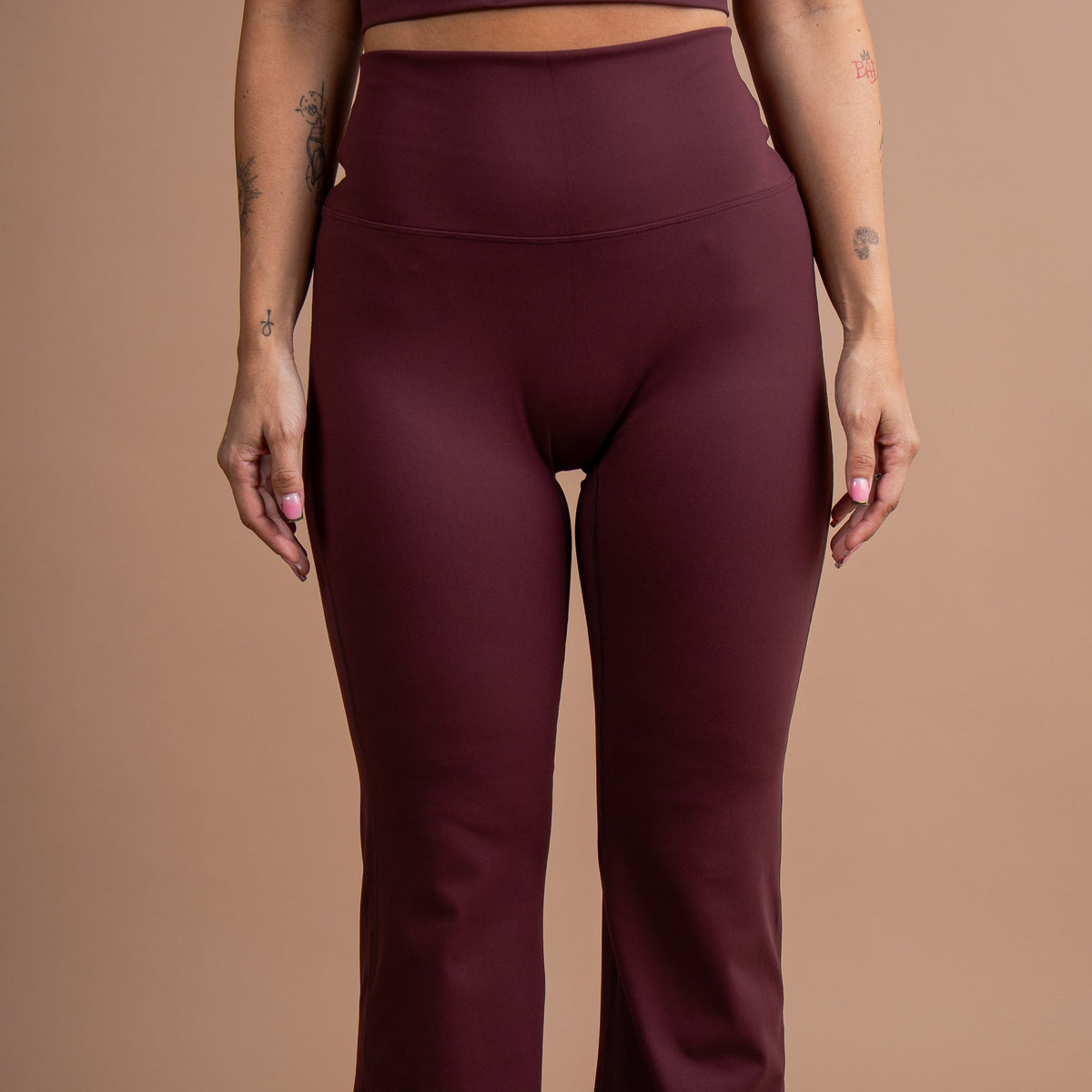 A model wearing a matching burgundy activewear set consisting of a scoop-neck crop top and high-waisted flared leggings, shown against a solid warm beige studio background.