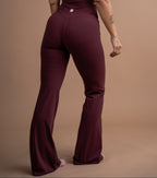 A woman wearing a deep burgundy one-piece jumpsuit, shown from the back against a solid tan studio backdrop.