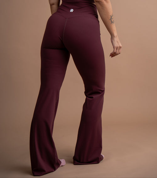 A woman wearing a deep burgundy one-piece jumpsuit, shown from the back against a solid tan studio backdrop.