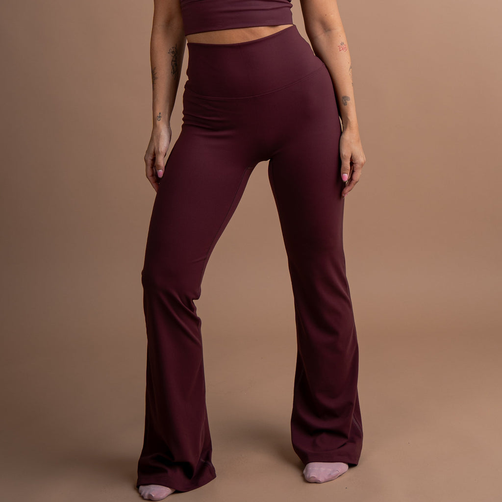 A woman modeling a matching burgundy activewear set consisting of a scoop-neck crop top and high-waisted flared leggings, standing against a solid warm brown studio backdrop.