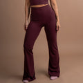 A woman modeling a matching burgundy activewear set consisting of a scoop-neck crop top and high-waisted flared leggings, standing against a solid warm brown studio backdrop.