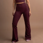 A woman modeling a matching burgundy activewear set consisting of a scoop-neck crop top and high-waisted flared leggings, standing against a solid warm brown studio backdrop.