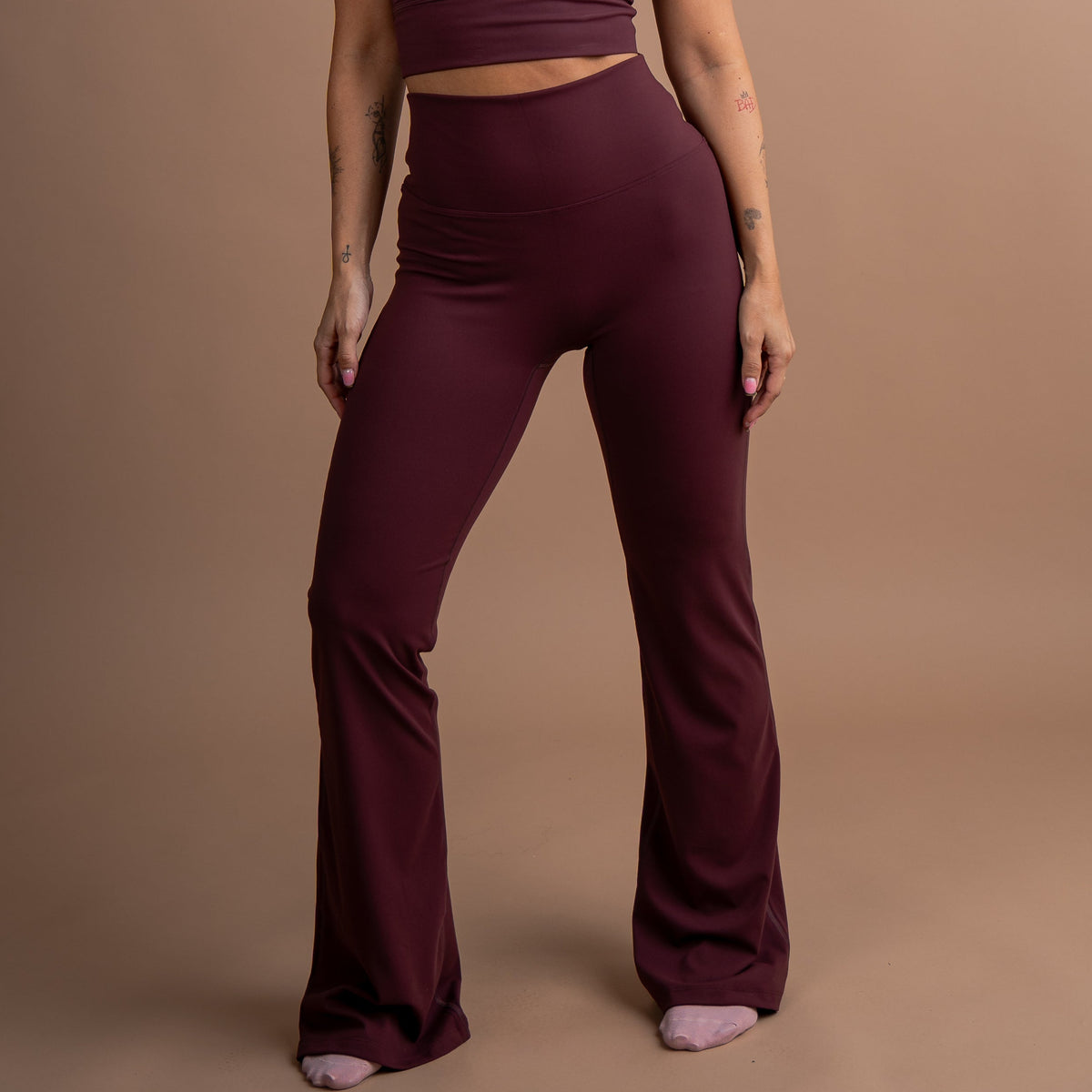 A woman modeling a matching burgundy activewear set consisting of a scoop-neck crop top and high-waisted flared leggings, standing against a solid warm brown studio backdrop.