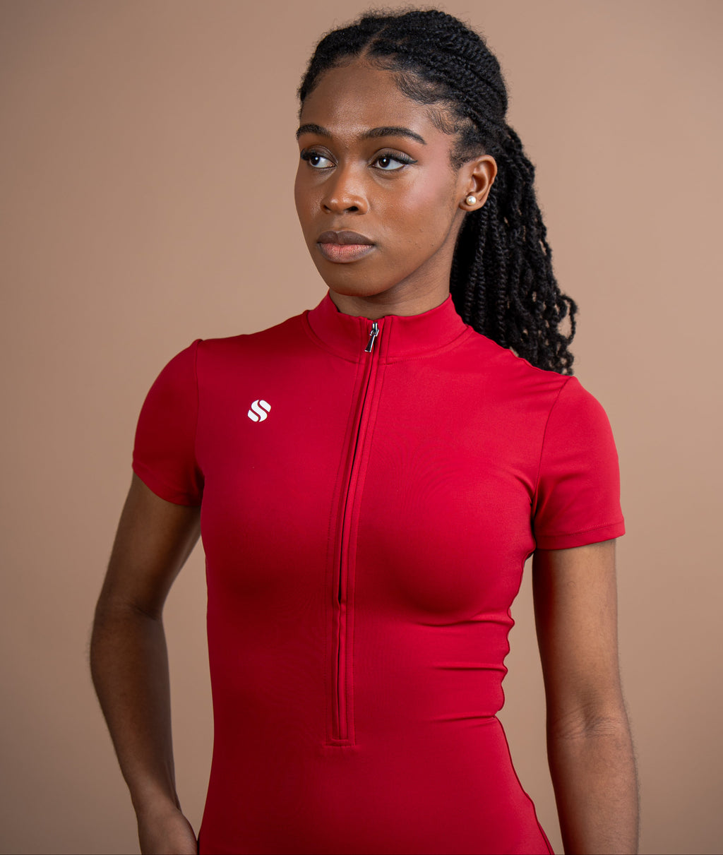 A woman wearing a bright red short-sleeve cycling jersey with a front zipper and a small white logo on the chest, photographed against a solid warm beige studio background.