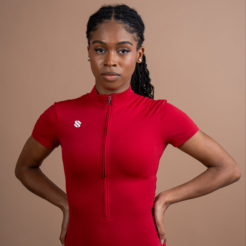A woman modeling a bright red short-sleeve zip-up athletic bodysuit against a solid warm brown studio background.