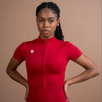 A woman modeling a bright red short-sleeve zip-up athletic bodysuit against a solid warm brown studio background.