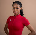 A woman with braided hair wearing a bright red short-sleeve cycling jersey with a front zipper, standing against a solid warm beige background.