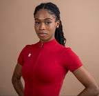 A woman with braided hair wearing a bright red short-sleeve cycling jersey with a front zipper, standing against a solid warm beige background.