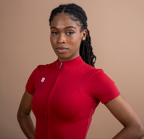 A woman with braided hair wearing a bright red short-sleeve cycling jersey with a front zipper, standing against a solid warm beige background.