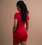 A woman with long black braids is shown from behind wearing a bright red short-sleeve athletic jumpsuit with matching biker shorts, set against a solid warm beige studio background.