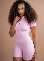 A woman with long, curly dark hair wearing a light pink short-sleeve one-piece bodysuit with a half-zip front, posed against a solid warm beige studio background.