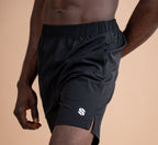A close-up of a man wearing black athletic shorts with a white logo on the lower left leg, set against a solid warm beige background.
