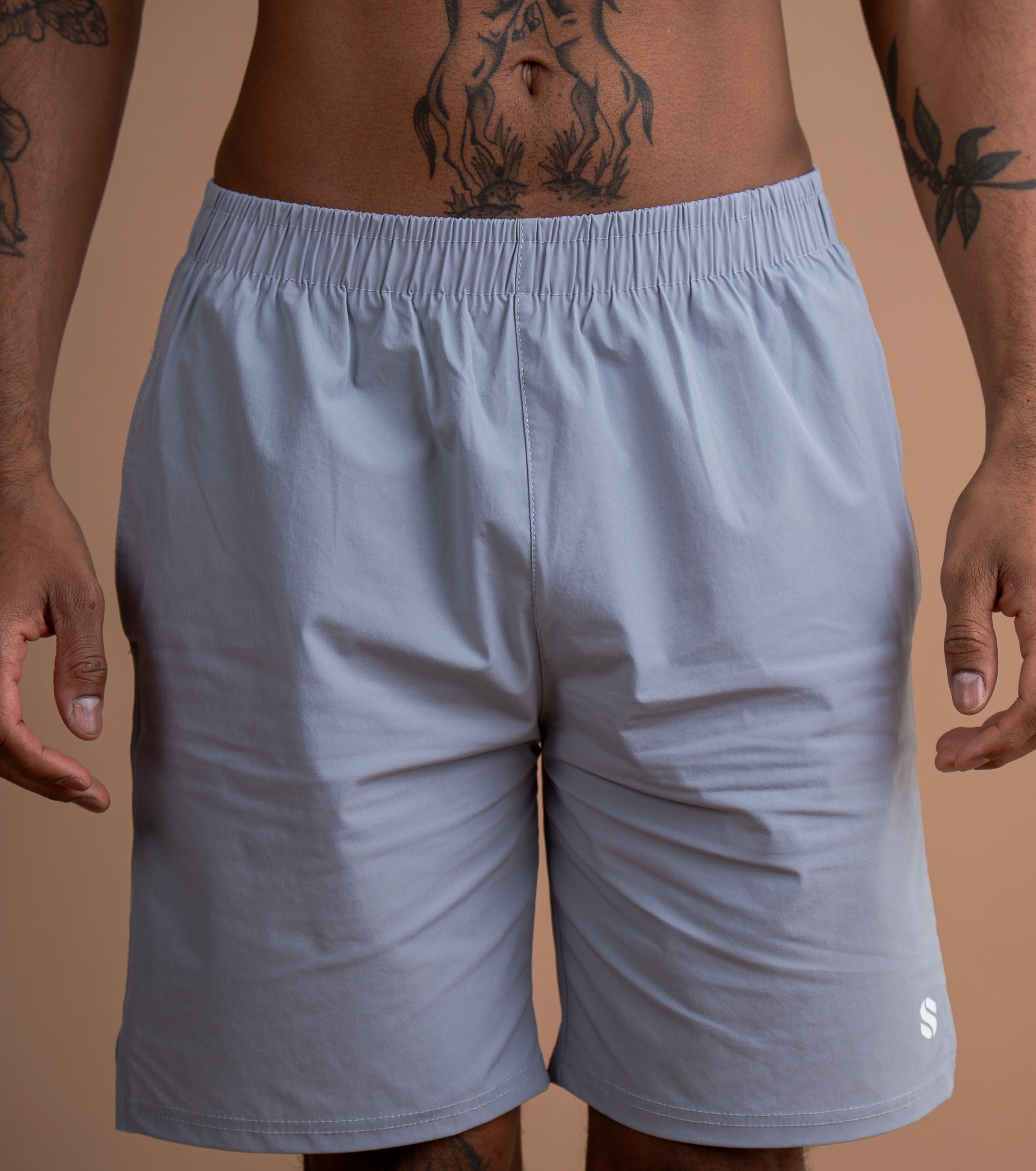 A close-up of a man wearing light gray athletic shorts, showing the waist, hips, and upper thighs. The man has visible tattoos on his torso and arms.