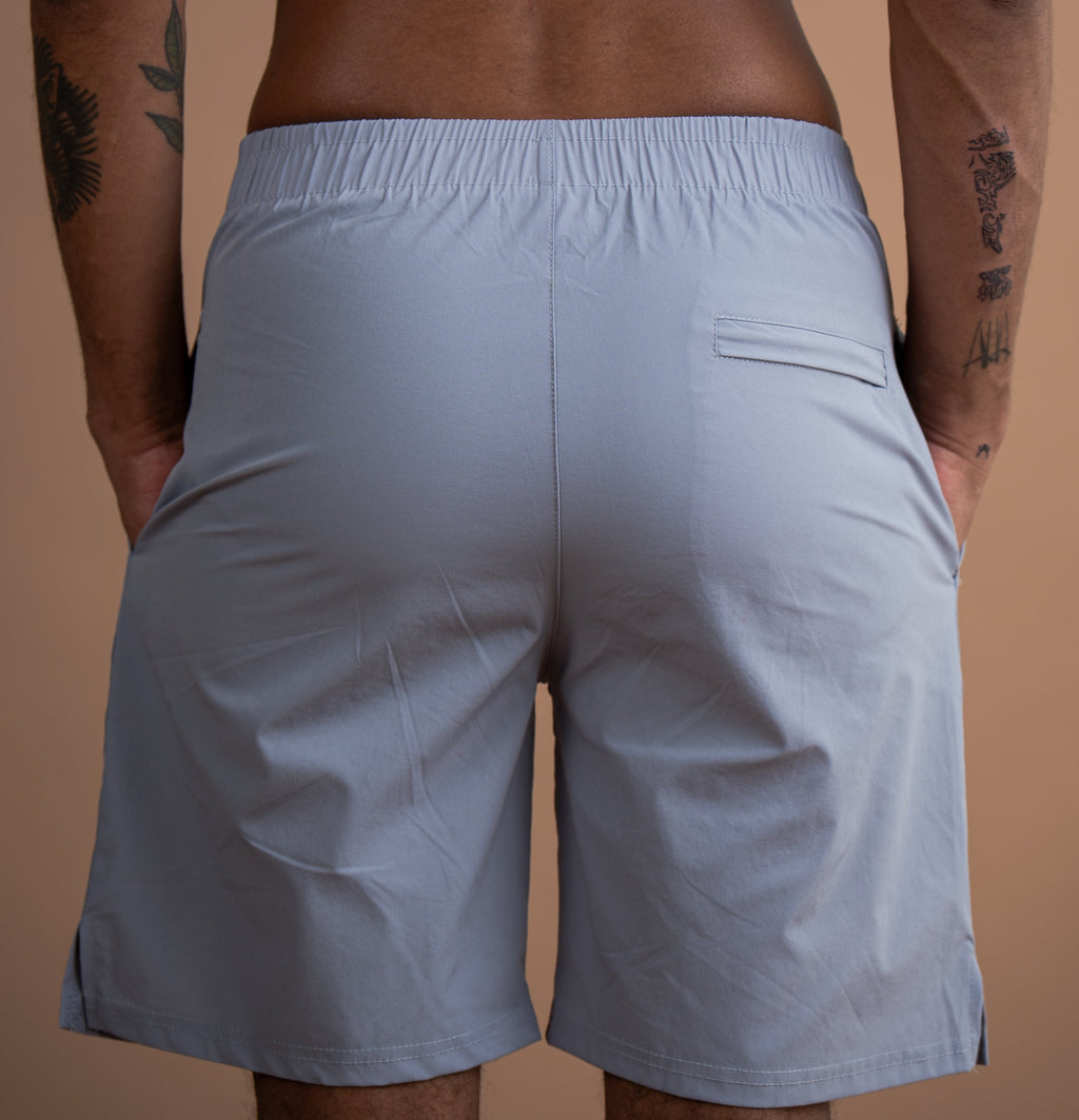 A man viewed from behind wearing light gray athletic shorts, showing the back waistband, side pocket, and leg fit against a plain tan background.
