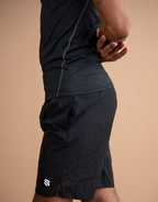 A close-up side view of a person wearing black athletic shorts and a matching black top against a solid warm beige studio background. A small white logo is visible on the lower left hem of the shorts.