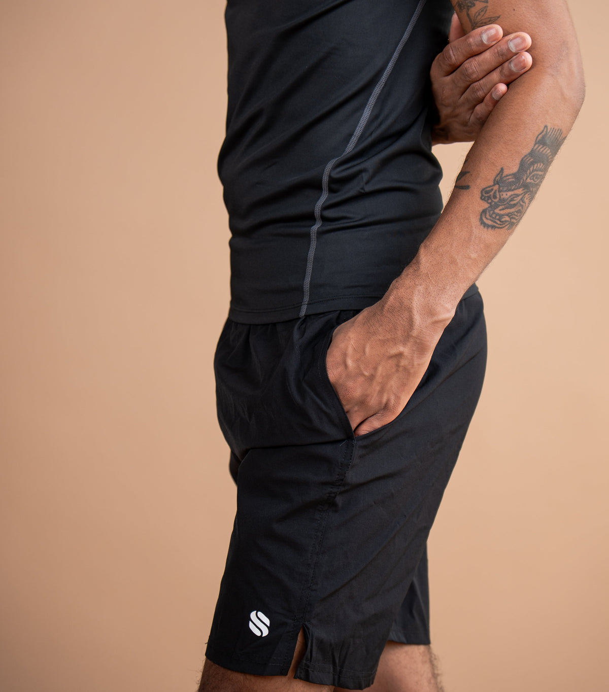 A man wearing a black athletic tank top and matching black shorts with a small white logo near the hem, posed against a solid tan studio background.