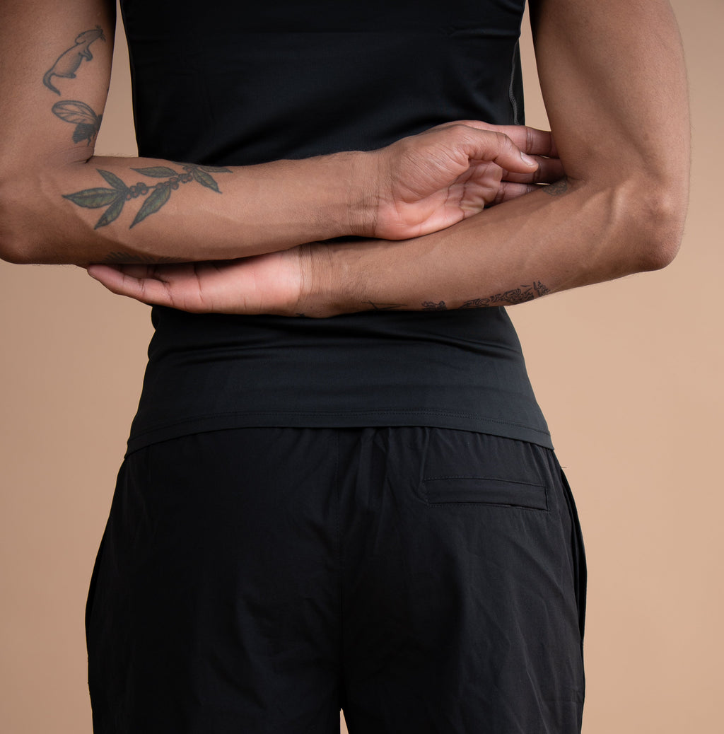 A person shown from the back wearing black athletic shorts and a black sleeveless top against a plain warm beige studio background.