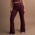 A woman modeling a matching burgundy activewear set consisting of a scoop-neck crop top and high-waisted flared leggings, standing against a solid warm brown studio backdrop.