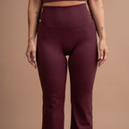 A model wearing a matching burgundy activewear set consisting of a scoop-neck crop top and high-waisted flared leggings, shown against a solid warm beige studio background.
