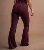 A woman wearing a deep burgundy one-piece jumpsuit, shown from the back against a solid tan studio backdrop.