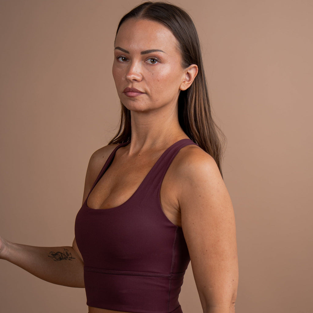 A woman modeling a matching burgundy activewear set, featuring a scoop-neck crop top and high-waisted leggings, set against a solid warm brown studio background.