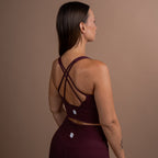 A woman shown from the back wearing a matching burgundy activewear set, featuring a sports bra with crisscross straps and high-waisted leggings, set against a solid taupe background.