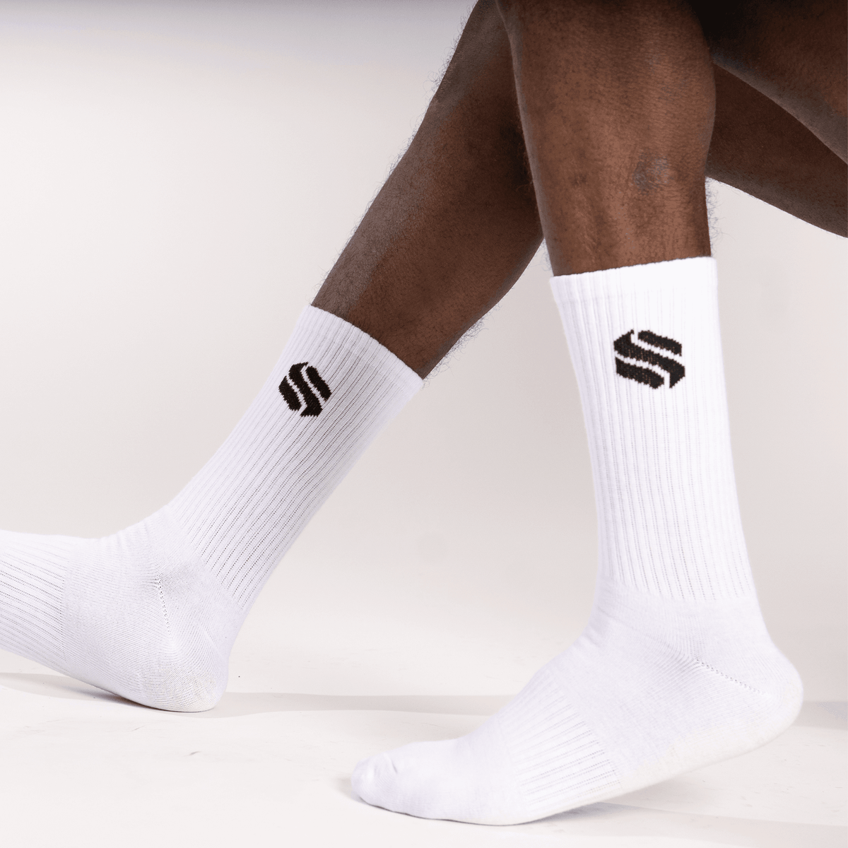 A pair of white crew-length socks on a person's legs, each featuring a black geometric S-shaped logo on the outer calf.
