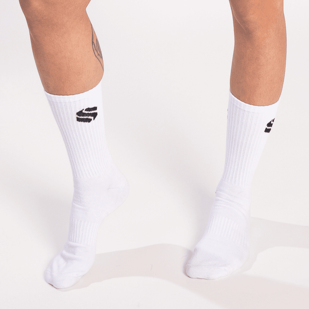 A pair of white crew-length socks worn on legs, each featuring a small black stylized letter logo on the outer calf.