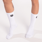 A pair of white crew-length socks worn on legs, each featuring a small black stylized letter logo on the outer calf.