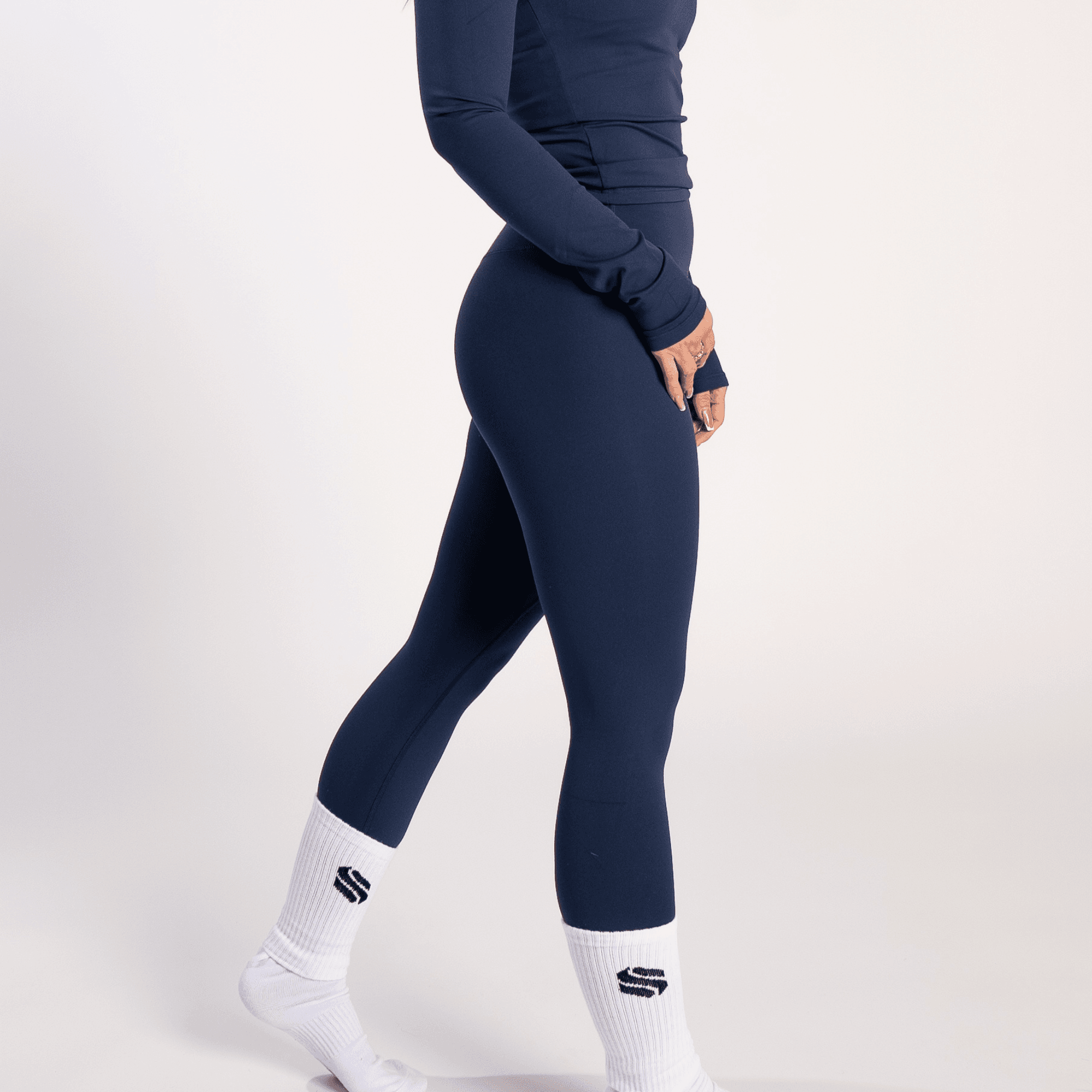 A person wearing navy blue leggings and a matching long-sleeved top with white mid-calf socks featuring a black logo on a plain white background.