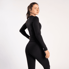 A woman stands sideways against a light grey background wearing a long-sleeved black fitted top and matching black leggings.