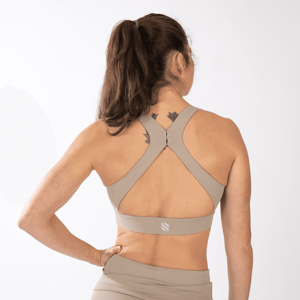 A beige sports bra with a racerback strap design and a small logo on the back band worn by a person from behind.